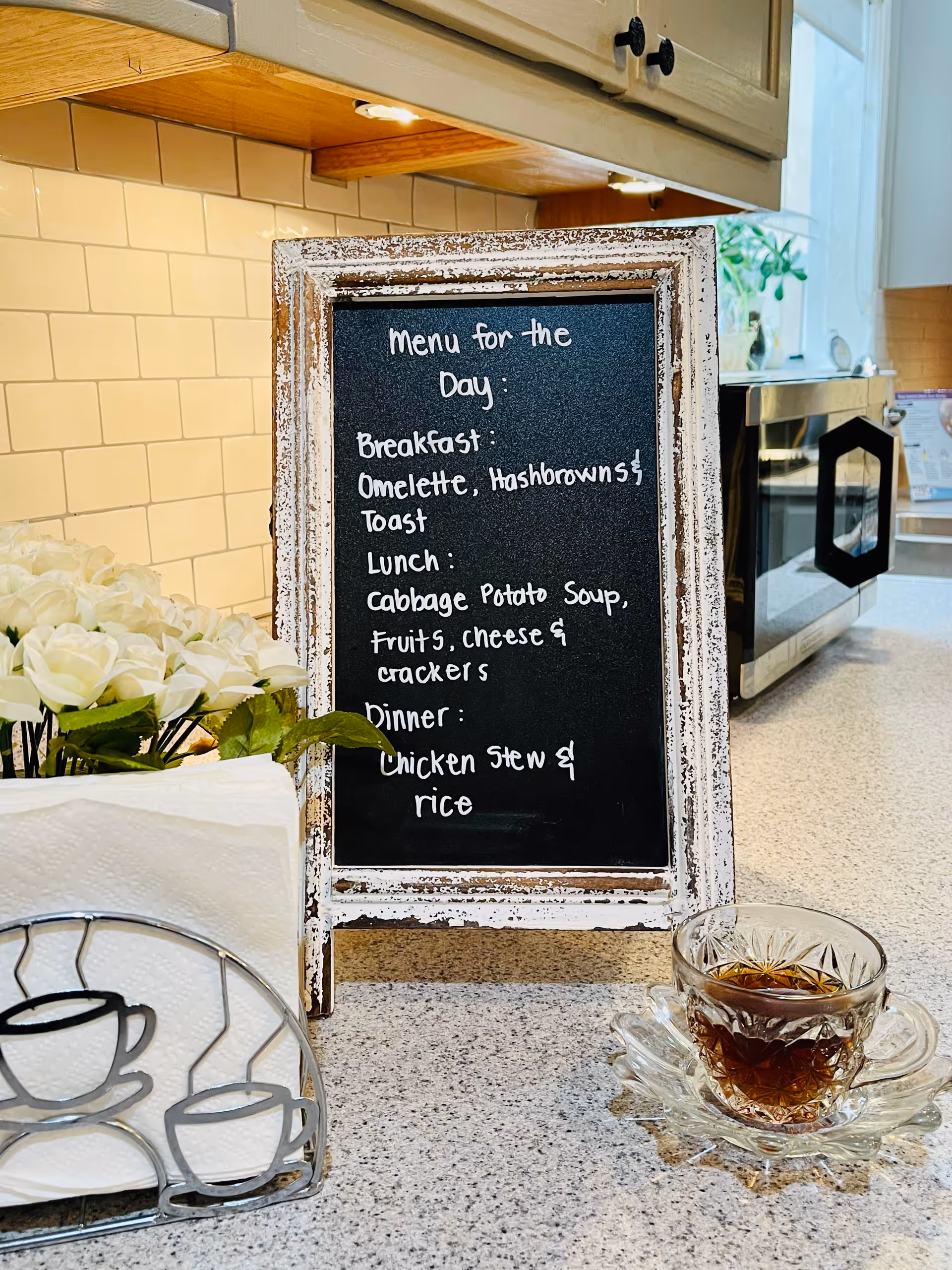 A kitchen countertop with a small chalkboard menu listing breakfast, lunch, and dinner items. The menu includes omelette, hashbrowns, toast for breakfast; cabbage potato soup, fruits, cheese, and crackers for lunch; and chicken stew with rice for dinner. Next to the menu is a glass cup with a dark beverage on a decorative glass saucer, a napkin holder with white napkins and a metal design of coffee cups, and a bunch of white artificial flowers. In the background, there is a microwave and kitchen cabinets with under-cabinet lighting.