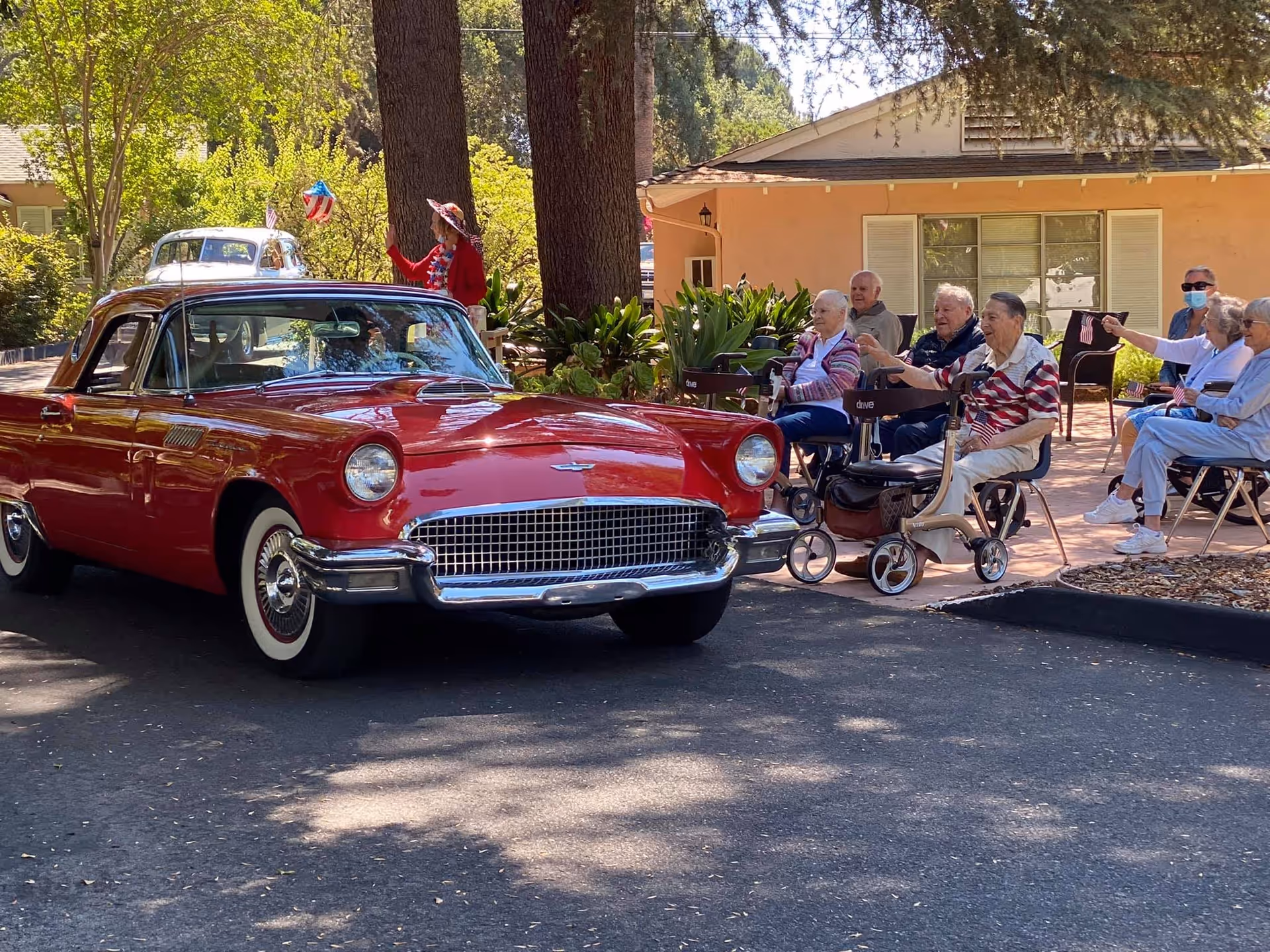 A group of elderly people sitting outdoors on chairs and mobility scooters, watching a shiny red vintage car parked on the road near a building with peach-colored walls and white window shutters. Trees and greenery surround the area, and a person in a red outfit and hat is standing near the trees in the background.