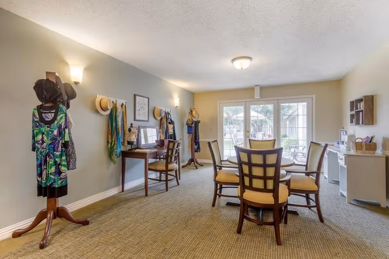 A well-lit room with a round table surrounded by five wooden chairs with yellow cushions. The room has a carpeted floor and light-colored walls. On the left side, there are coat racks with hats, scarves, and a dress displayed. A small wooden desk with a chair is also visible along the left wall. At the far end, large glass doors open to an outdoor area with patio furniture. On the right side, there is a white desk with baskets and some items on top, along with a small wall-mounted shelf.