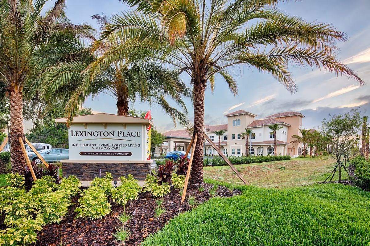 Entrance sign for Lexington Place senior living community surrounded by palm trees and landscaping, with the facility building visible in the background under a partly cloudy sky.