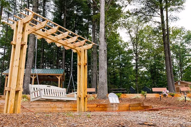 A wooden swing bench hanging from a wooden pergola structure in an outdoor garden area surrounded by tall trees. There are benches, a small covered shelter, and a flower arrangement in the background.