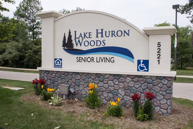 Outdoor sign for Lake Huron Woods Senior Living with the address number 5221, surrounded by flowers and greenery.