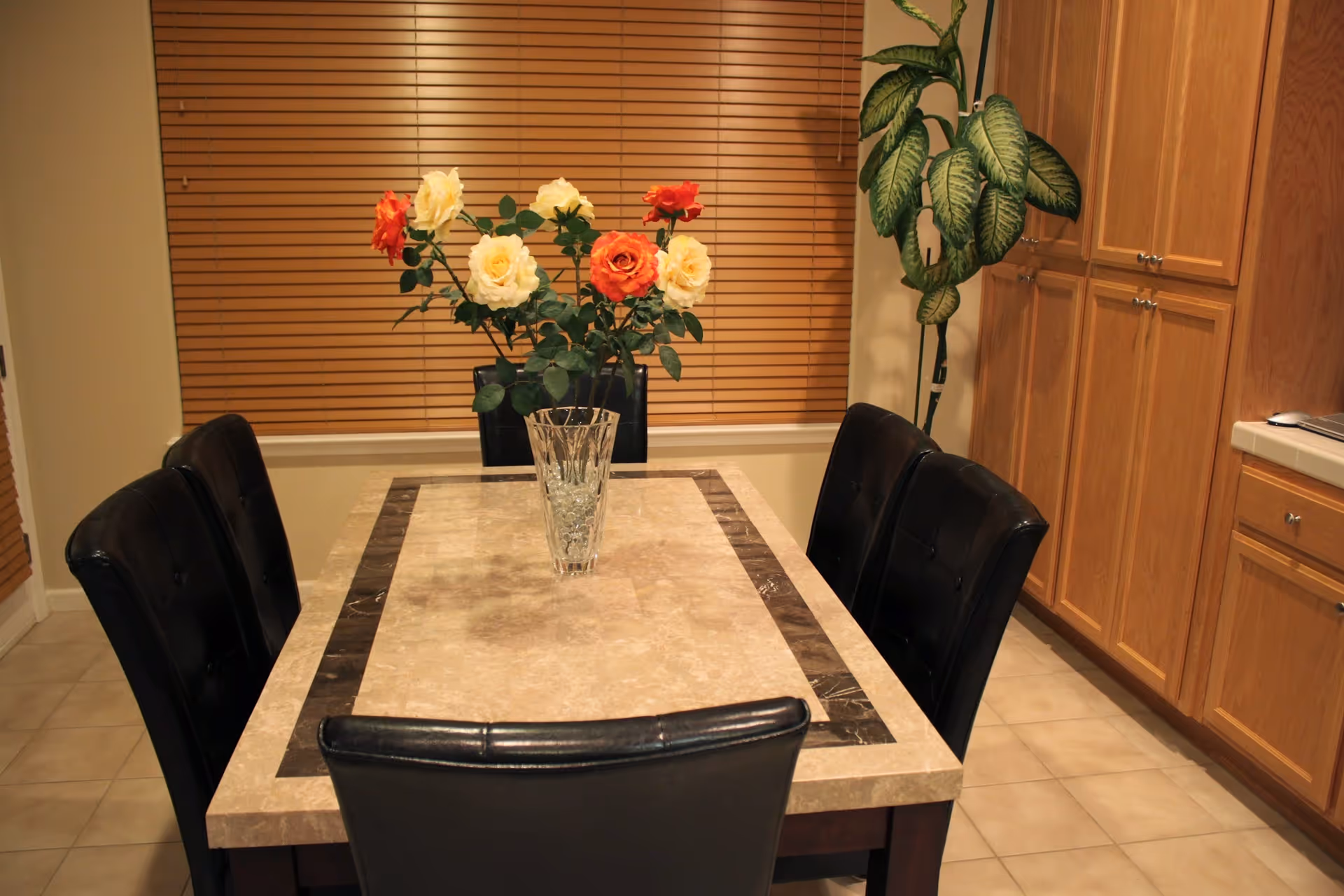 A dining area with a rectangular marble table surrounded by six black leather chairs. A glass vase with yellow and orange roses is placed in the center of the table. Behind the table, there is a window with closed wooden blinds and a tall green potted plant to the right. Wooden cabinets and tiled floor are also visible.