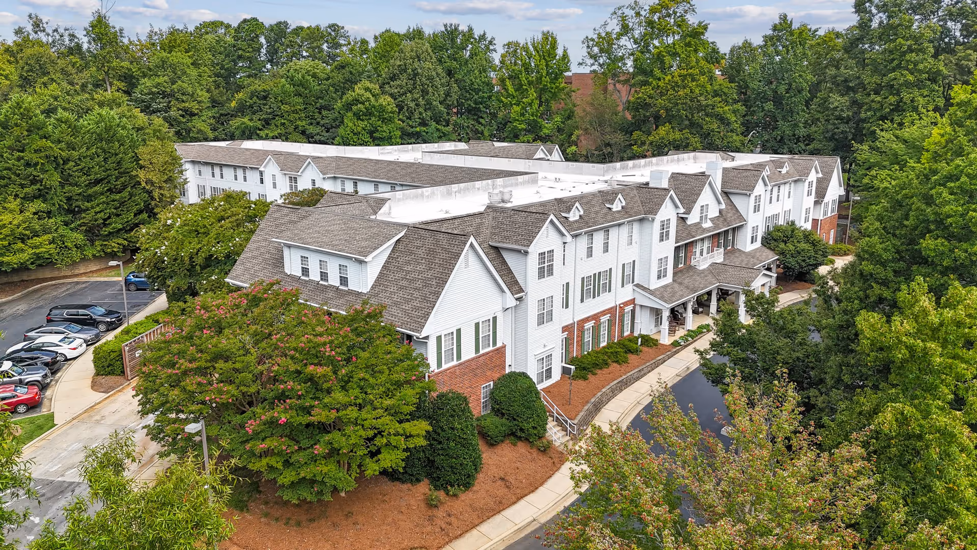 Aerial view of a large senior living facility building surrounded by lush green trees and landscaping. The building has white siding with brick accents and multiple gabled roofs. There is a curved driveway and parking area with several cars parked. The sky is partly cloudy.