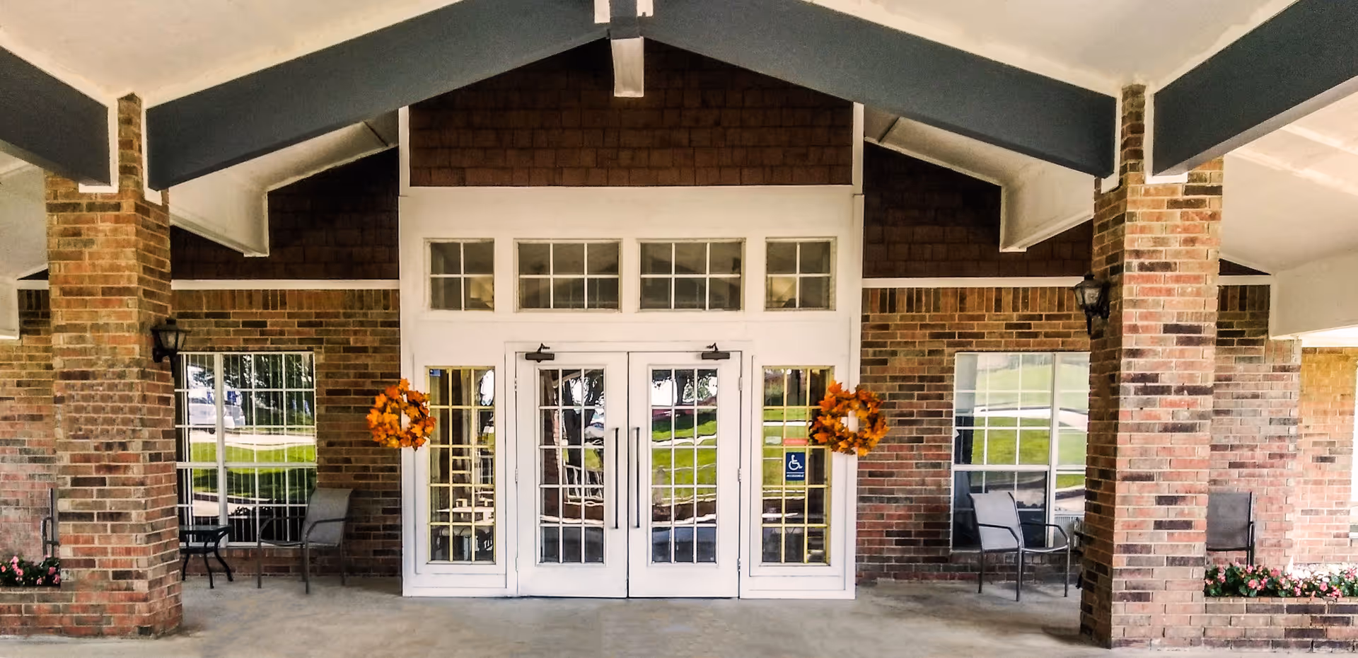 Entrance of a brick building with white double doors and windows above. The entrance is decorated with two orange wreaths on either side of the doors. There are brick columns supporting a covered porch area with seating visible through the windows.