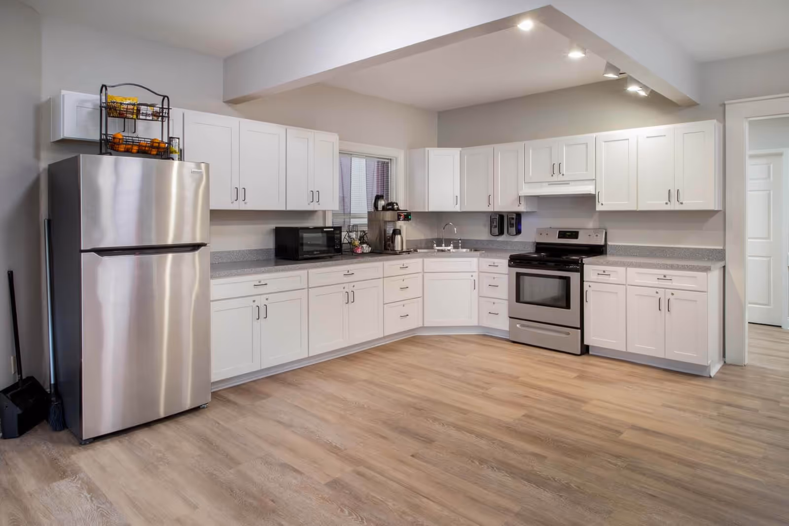 A modern kitchen with white cabinets, a stainless steel refrigerator, a black microwave, a coffee maker, a stainless steel stove with an oven, and a double sink. The kitchen has light wood flooring and gray countertops, with overhead lighting and a window above the sink.