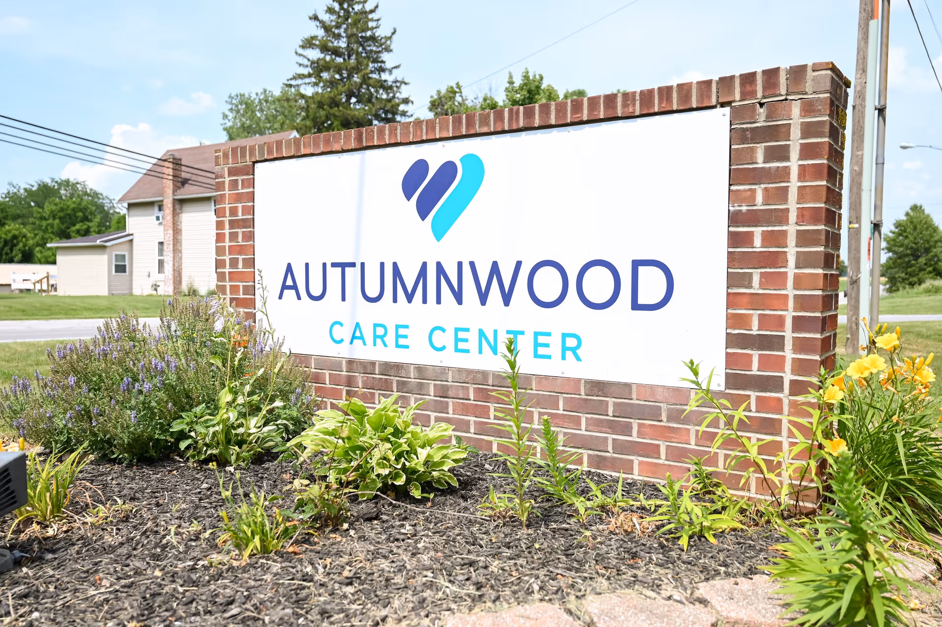 A brick sign for Autumnwood Care Center surrounded by plants and flowers, with a clear blue sky and some buildings in the background.
