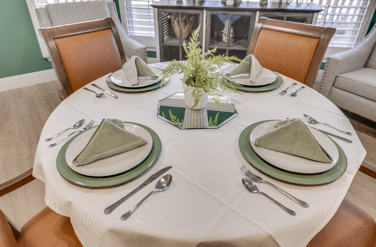 A round dining table set for four with white tablecloth, green placemats, white plates, green folded napkins, and silver cutlery. A small potted fern centerpiece sits on a mirrored tray in the middle of the table. The background shows wooden chairs with brown leather seats and a cabinet with glass doors.