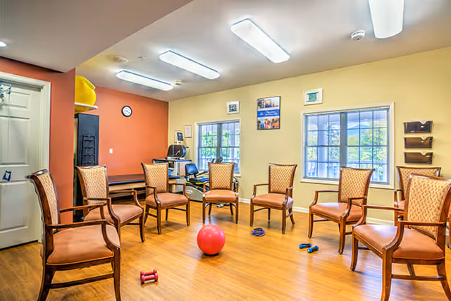 A bright room with wooden flooring and several wooden chairs arranged in a circle. There are exercise equipment items such as small dumbbells and a red exercise ball on the floor. The walls are painted in warm tones with windows letting in natural light. A clock and some framed pictures are visible on the walls.