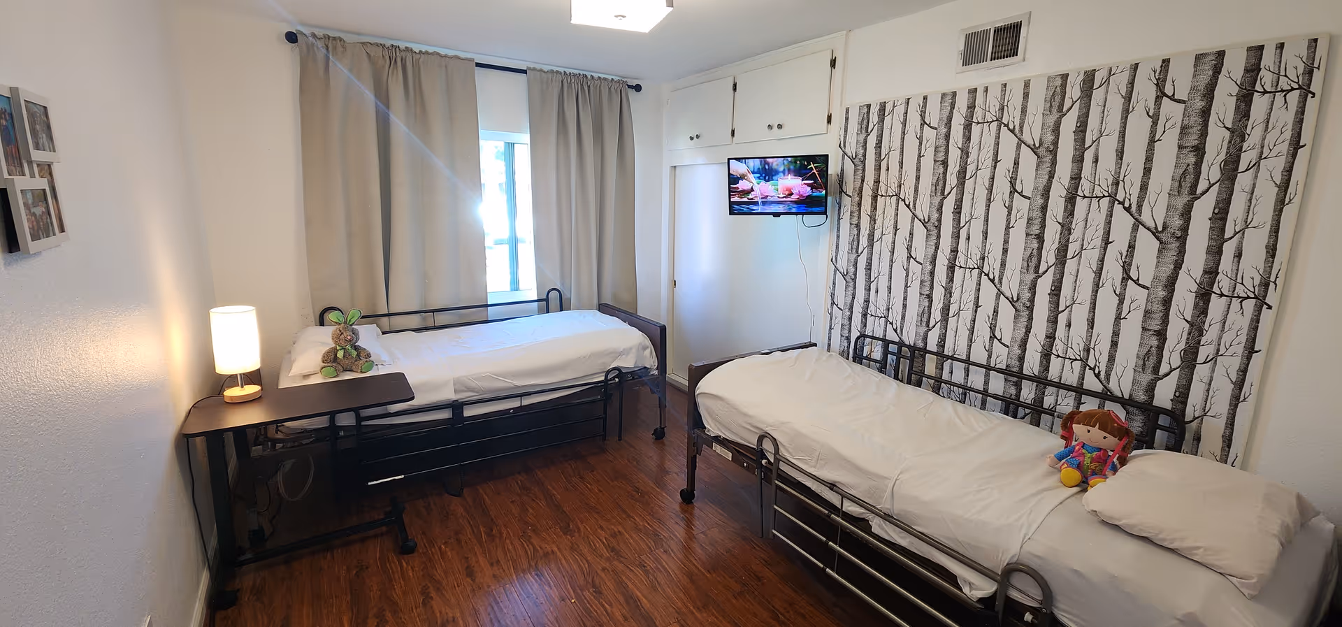 A small bedroom with two single beds, each with white bedding. One bed has a stuffed bunny toy and a small lamp on a bedside table, while the other bed has a doll placed on it. The room has wooden flooring, a window with beige curtains, a wall-mounted TV, and a wallpaper with a black and white tree pattern on one wall.