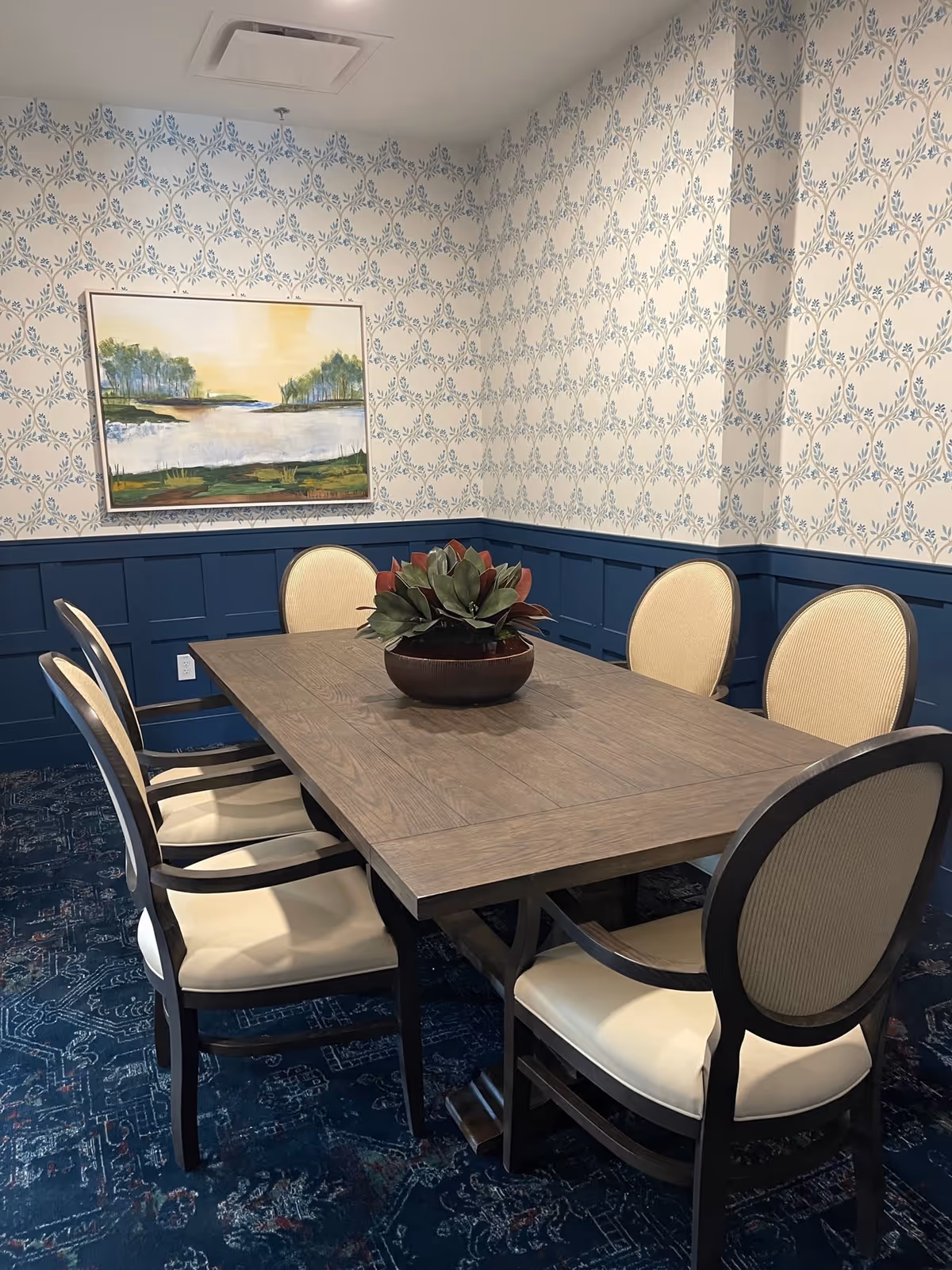 A small dining room with a rectangular wooden table surrounded by six upholstered chairs. The walls have a blue and white patterned wallpaper with blue wainscoting. A framed landscape painting hangs on the wall above the table, and a potted plant centerpiece is on the table. The floor is covered with a dark blue patterned carpet.