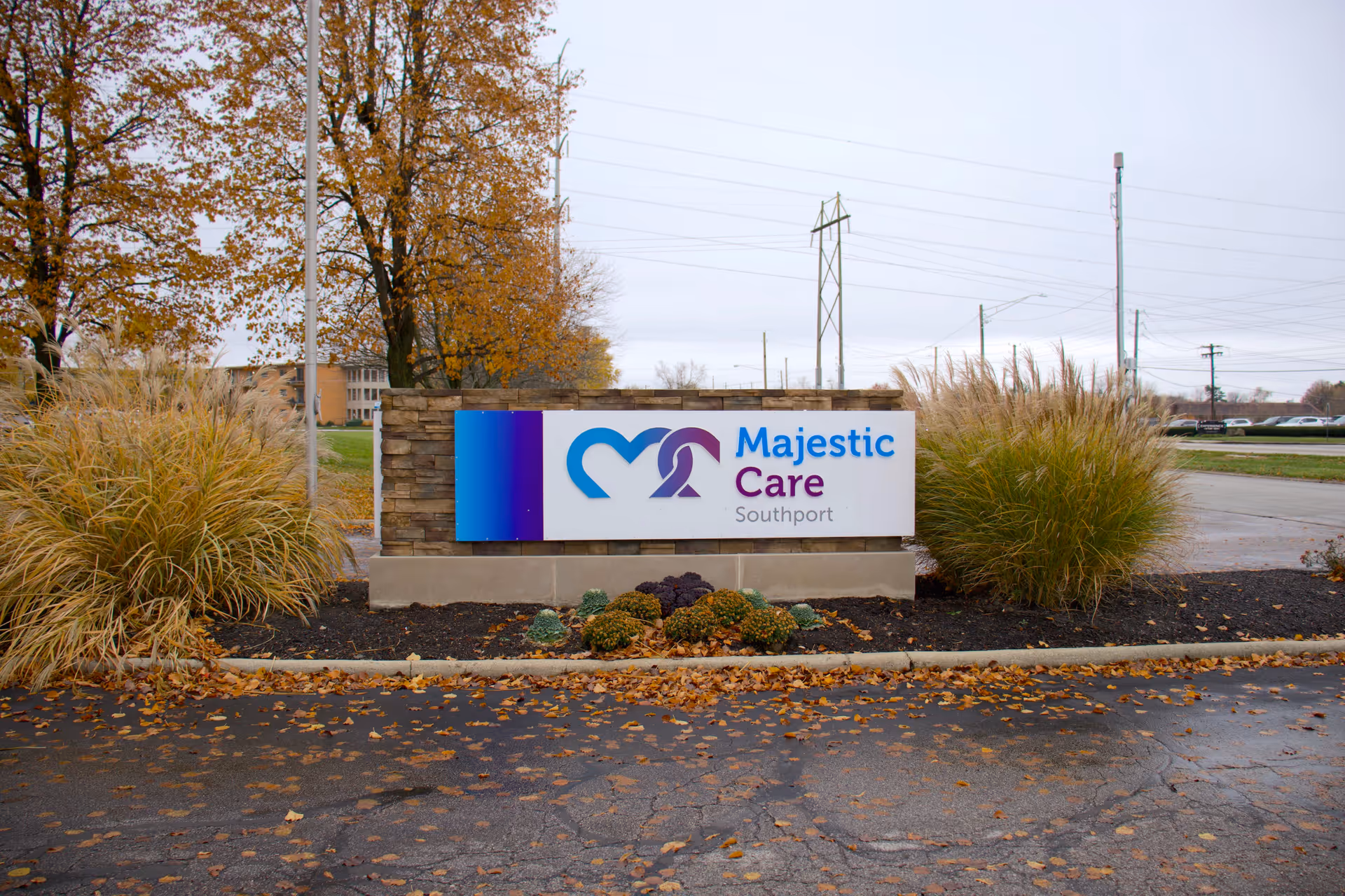 Outdoor view of a sign for Majestic Care Southport, surrounded by ornamental grasses and small shrubs, with autumn trees and a cloudy sky in the background.