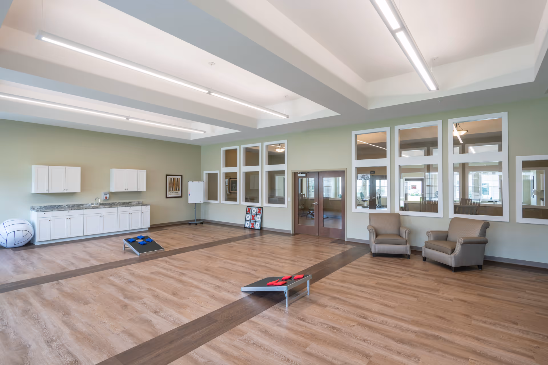 A spacious, well-lit activity room with light green walls and wood flooring. The room features two armchairs, a large tic-tac-toe game, a whiteboard, and a cabinet with a countertop along one wall. There are two cornhole boards with bean bags on the floor, and large windows and double doors leading to another room.