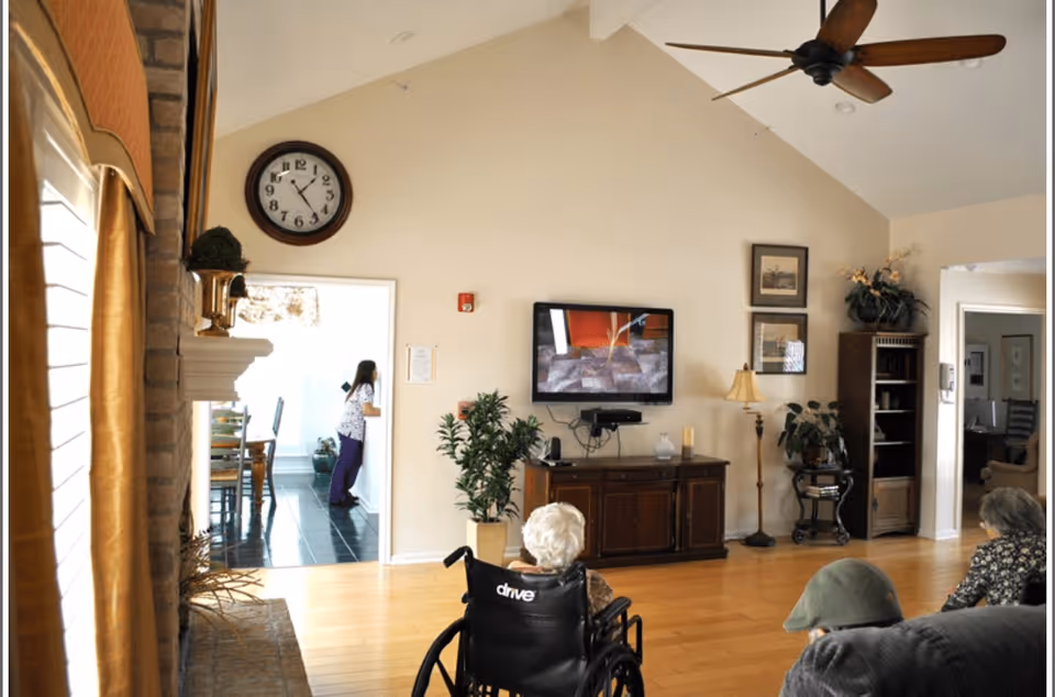 A living room in an assisted living facility with a high vaulted ceiling and a ceiling fan. An elderly person in a wheelchair is watching a wall-mounted TV. The room has wooden flooring, a wooden cabinet under the TV, framed pictures on the wall, a floor lamp, and several plants. A woman is seen in an adjacent room through a doorway, which appears to be a dining area with a table and chairs.