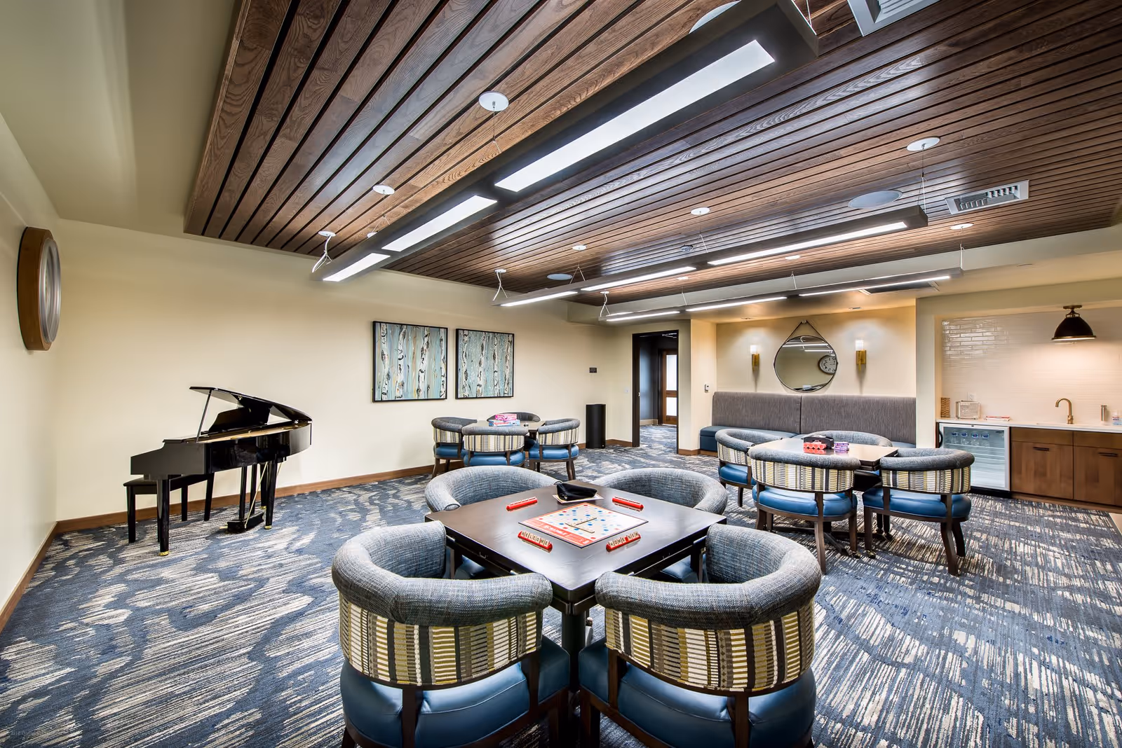 A spacious common room with a patterned carpet, wooden ceiling with modern lighting, and several round tables surrounded by cushioned chairs. A black grand piano is positioned against one wall, and there is a small kitchenette area with a sink, cabinets, and a mini fridge. The room has neutral-colored walls with decorative artwork and a round mirror above a cushioned bench.