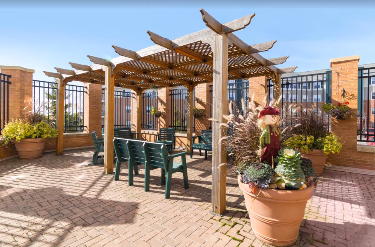Outdoor patio area with a wooden pergola providing partial shade over green benches and chairs. Large potted plants with decorative grasses and a small scarecrow figure are placed around the space. The area is enclosed by a brick and metal fence, with a building visible in the background under a clear blue sky.