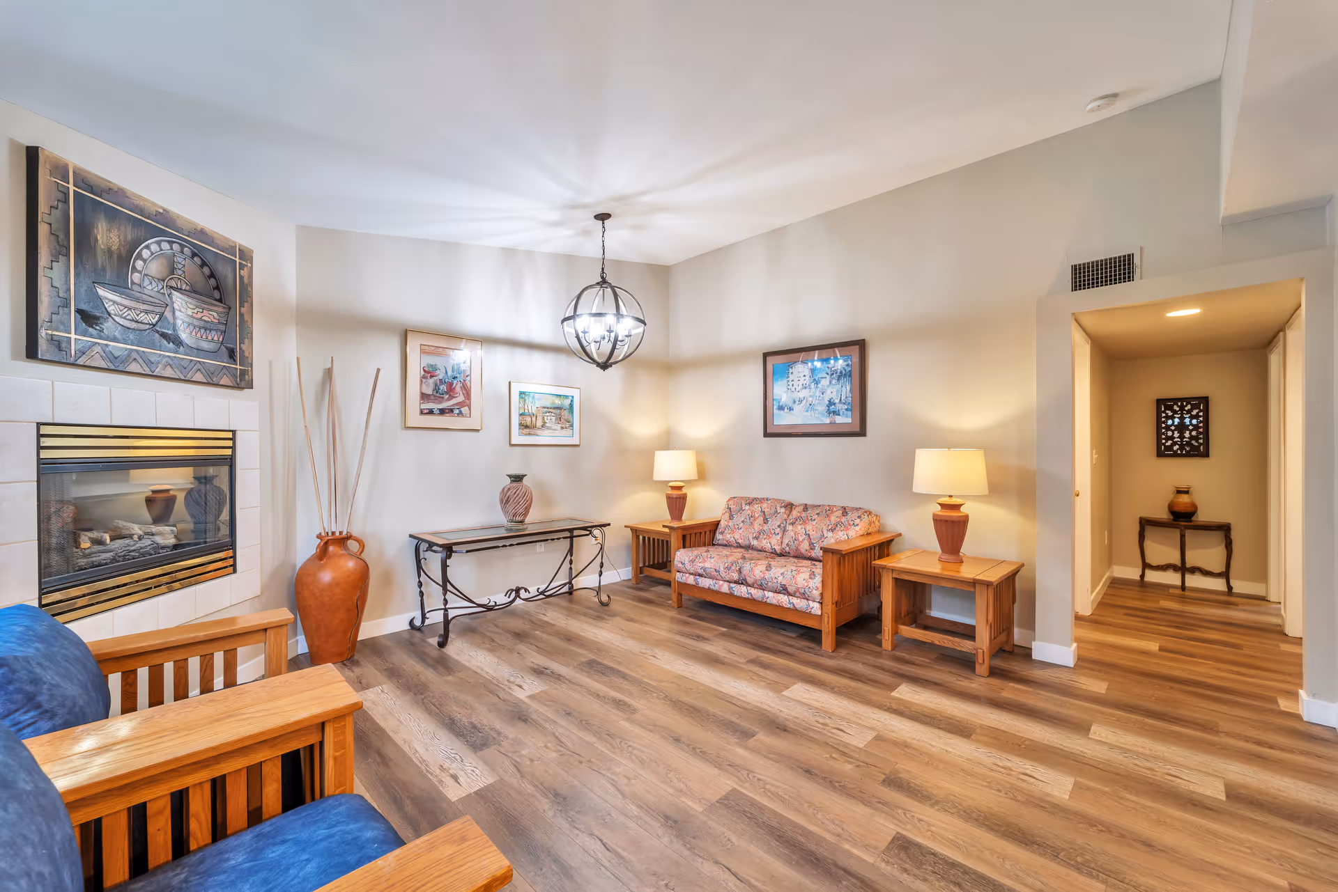 A cozy living room with wooden furniture including a floral-patterned couch, two side tables with lamps, and a blue cushioned chair. The room features a fireplace with decorative artwork above it, a metal and glass console table with a vase, and framed pictures on the walls. The floor is wood laminate, and a modern spherical chandelier hangs from the ceiling.