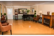 A cozy living room area with wooden flooring, a black grand piano, two brown leather chairs, a wooden electric fireplace, a floor lamp, and decorative plants. There is a door and windows letting in natural light.
