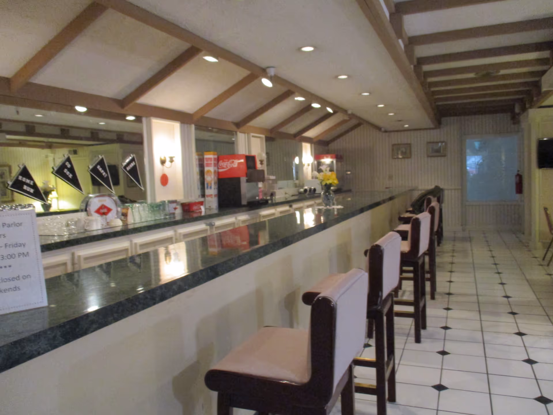 Interior view of a senior living facility's dining or snack bar area with a long counter, several high chairs with light pink cushions, a Coca-Cola soda dispenser, and a popcorn machine. The ceiling has exposed wooden beams and recessed lighting, and the floor is tiled with a white and black diamond pattern.