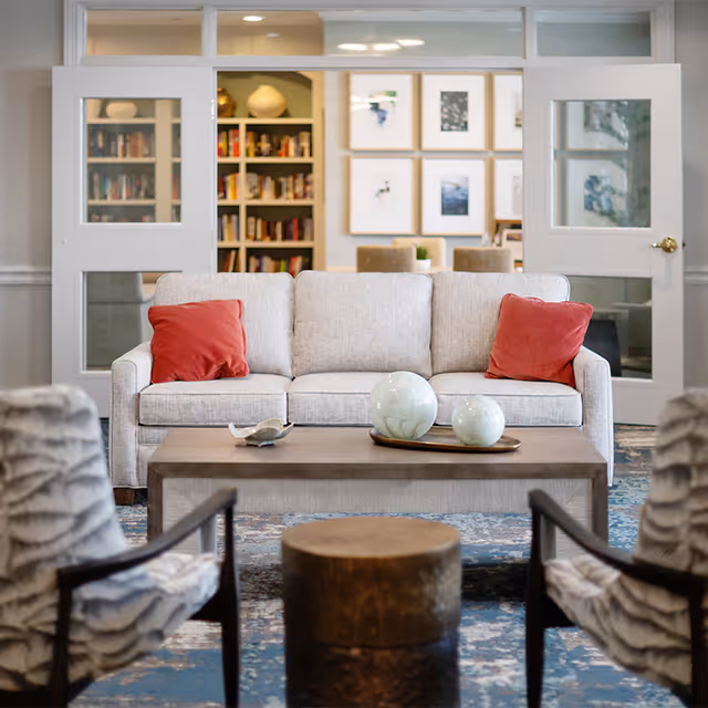 A cozy living room area with a light gray sofa adorned with two red throw pillows. In front of the sofa is a wooden coffee table with decorative ceramic spheres and a small dish. Two patterned armchairs face the sofa, and a round wooden stool is positioned between them. Behind the sofa, there are glass-paned double doors leading to a room with bookshelves and framed pictures on the wall.