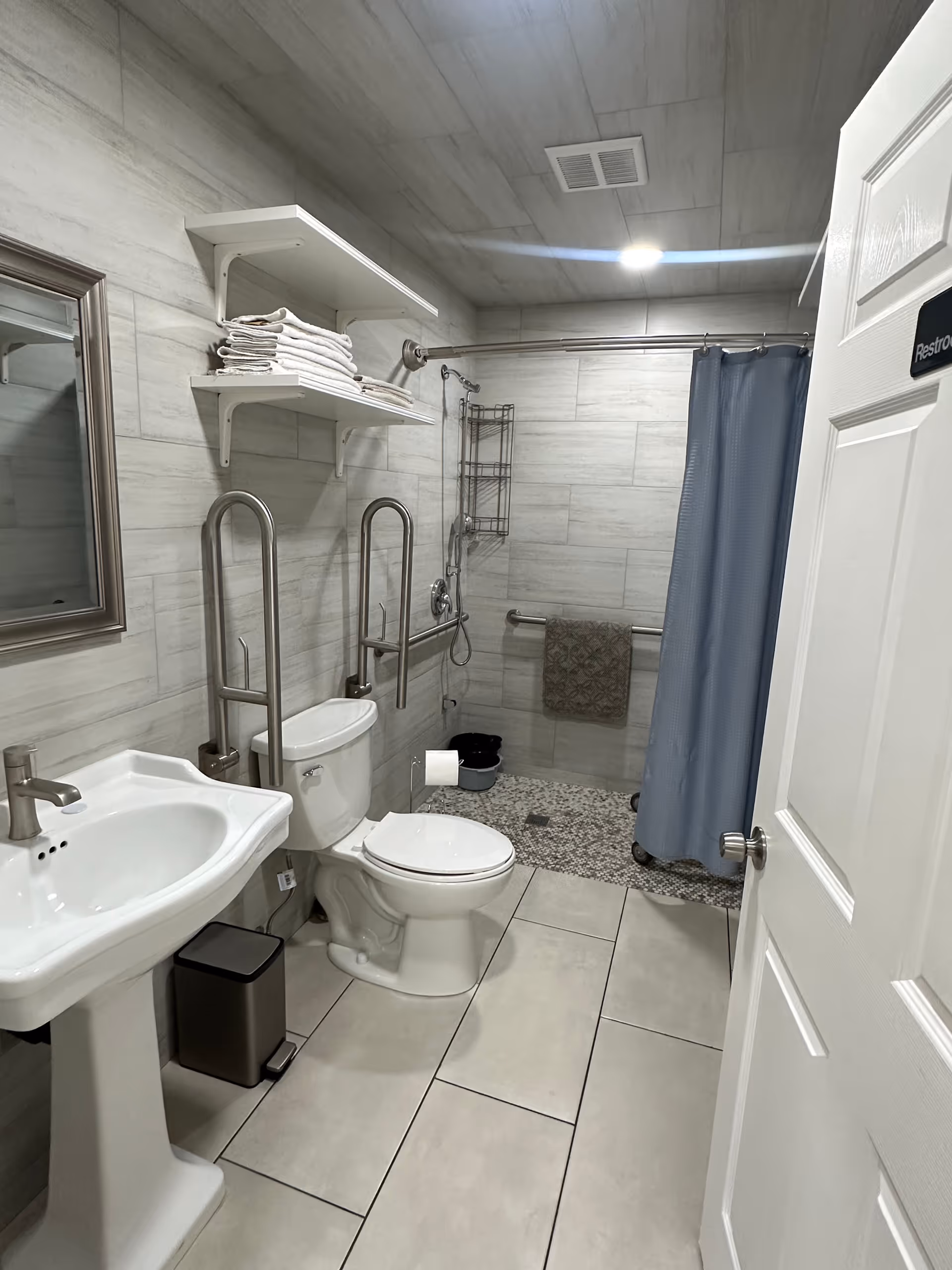A clean, accessible bathroom with a pedestal sink, a toilet equipped with grab bars, a wall-mounted mirror, and a walk-in shower with a blue curtain and grab bars. Shelves above the toilet hold neatly folded towels. The floor and walls are tiled in light colors, and a towel hangs on a bar inside the shower area.