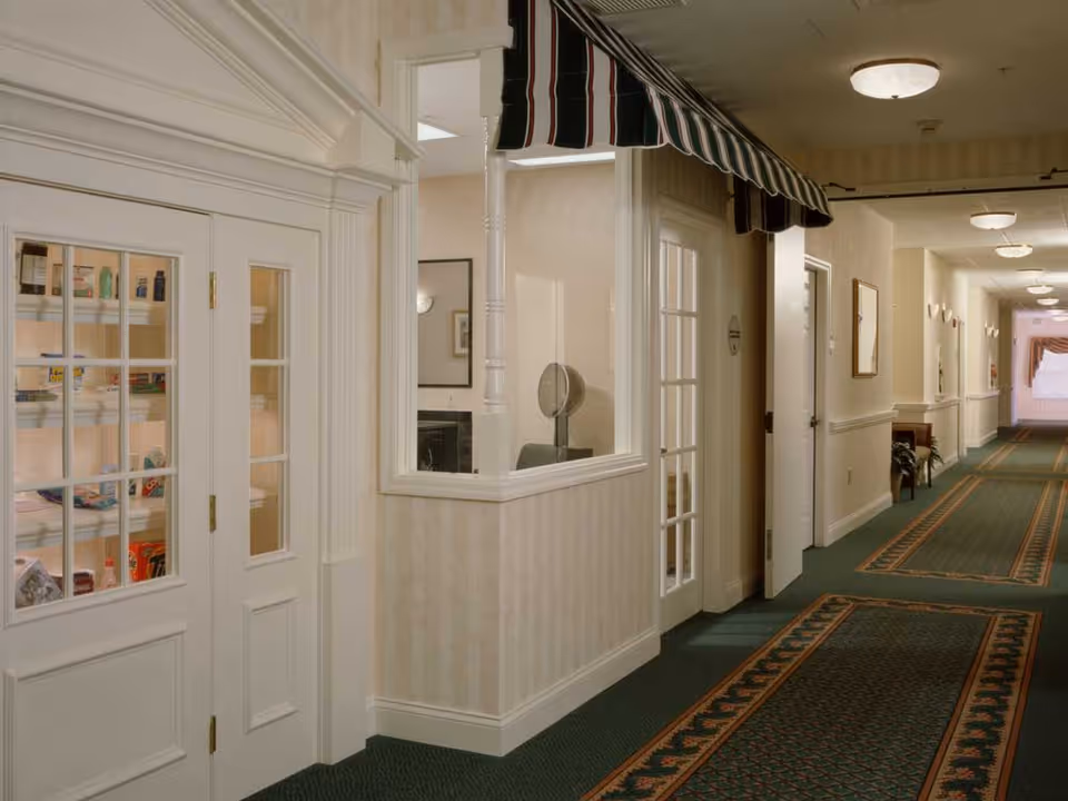 Interior hallway of a senior living facility with patterned green carpet and cream-colored walls. On the left, there is a small room with glass-paned double doors displaying shelves stocked with various items. Next to it is a window with a striped green and white awning above it. The hallway extends into the distance with multiple doors and framed pictures on the walls.