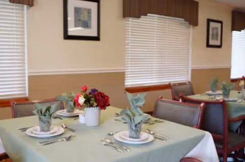 A dining room with tables set for meals, flowers, and window blinds.