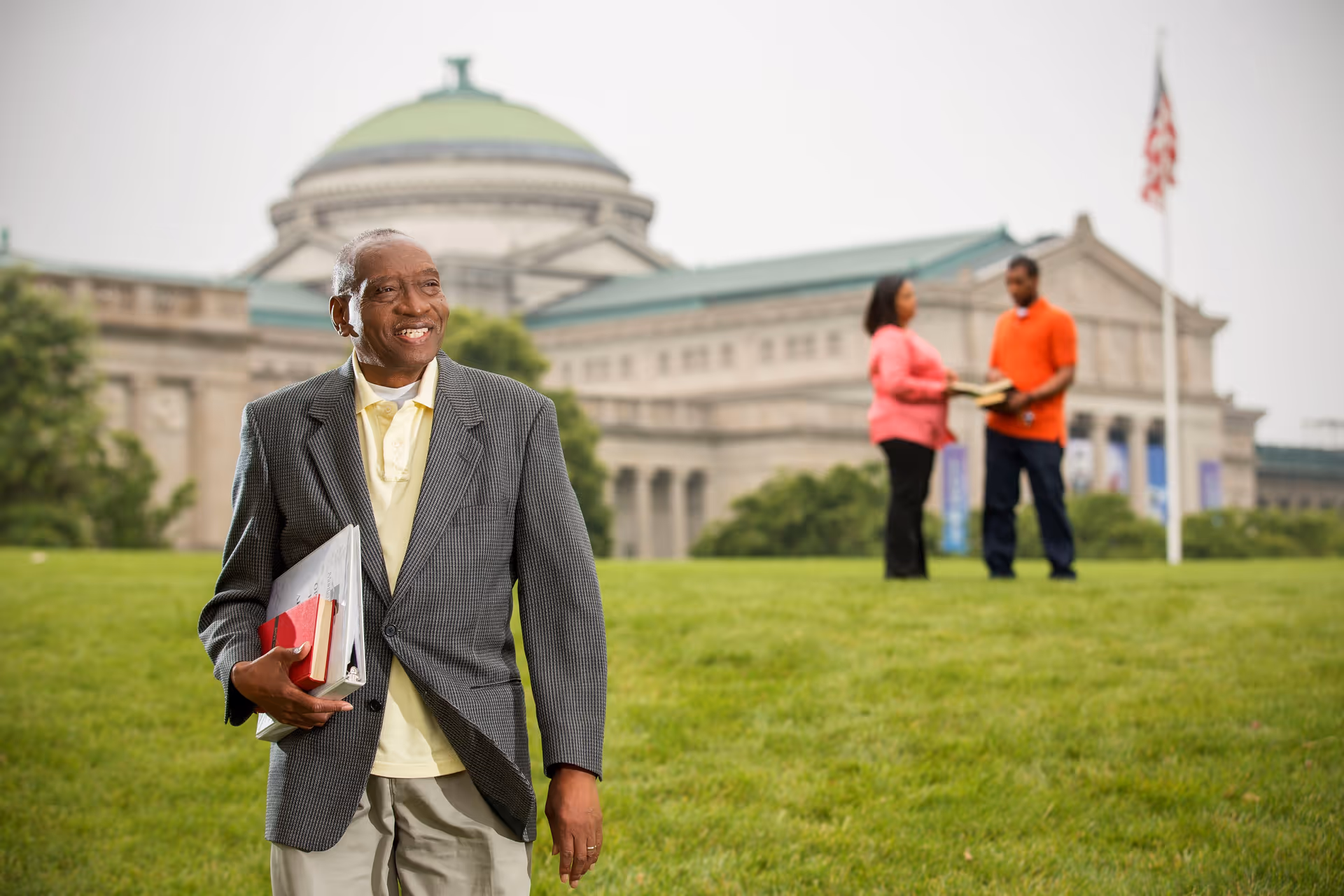 An older man wearing a gray blazer and holding books stands on a grassy area with a large domed building in the background. Two people, a woman in a pink top and a man in an orange shirt, are standing and talking in the distance near an American flag.