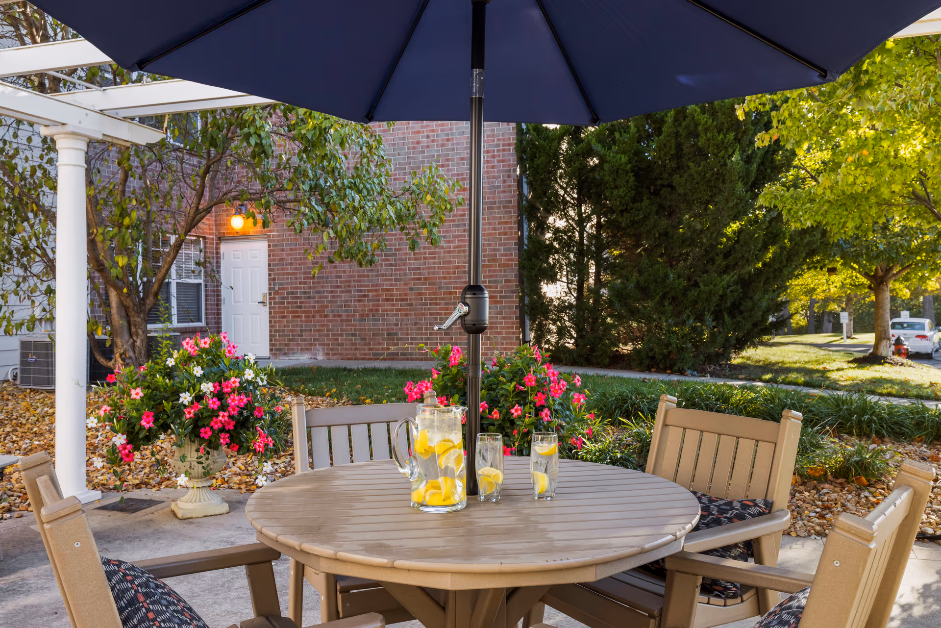 Outdoor patio area with a round wooden table and four chairs under a large blue umbrella. On the table, there is a pitcher and glasses filled with lemon water. Surrounding the patio are green trees, pink flowers in planters, and a brick building with a white door in the background.