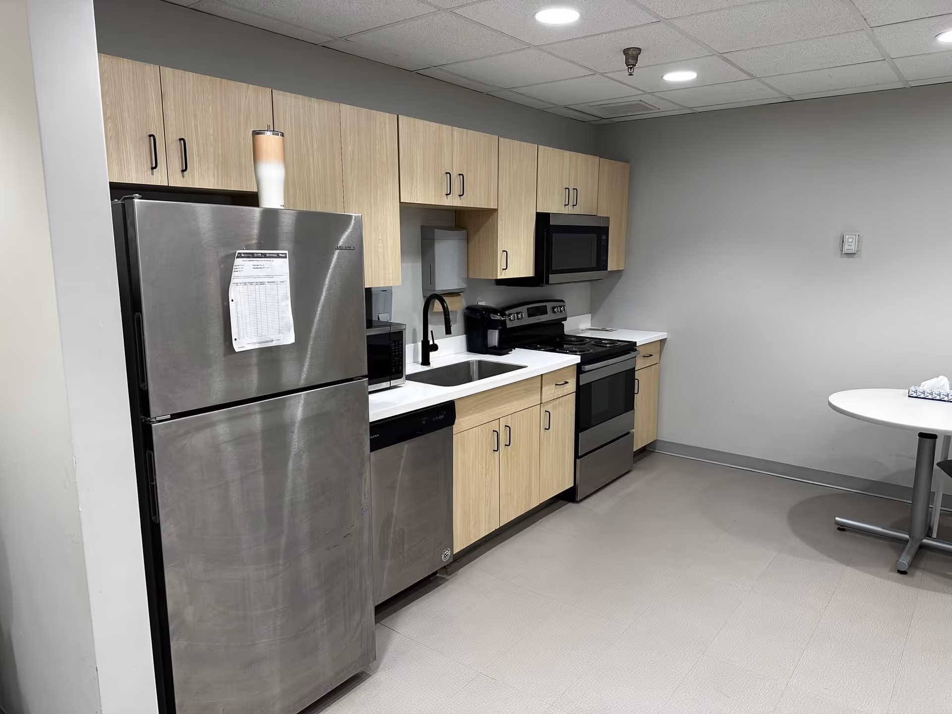 A modern kitchen area with light wood cabinets, a stainless steel refrigerator, dishwasher, oven, microwave, and a sink with a black faucet. There is a small round table with a tissue box on the right side against a plain gray wall.
