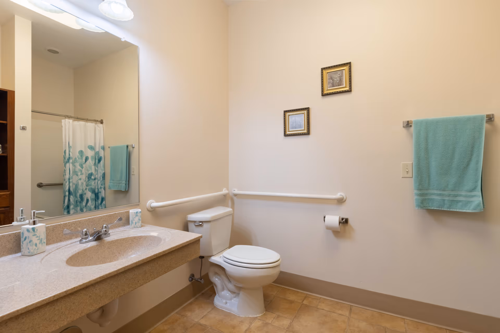 A clean and accessible bathroom with a beige countertop sink, a large mirror, a white toilet with grab bars on the walls, a shower with a curtain featuring a blue leaf pattern, and teal towels hanging on the wall. Two small framed pictures are hung above the toilet.