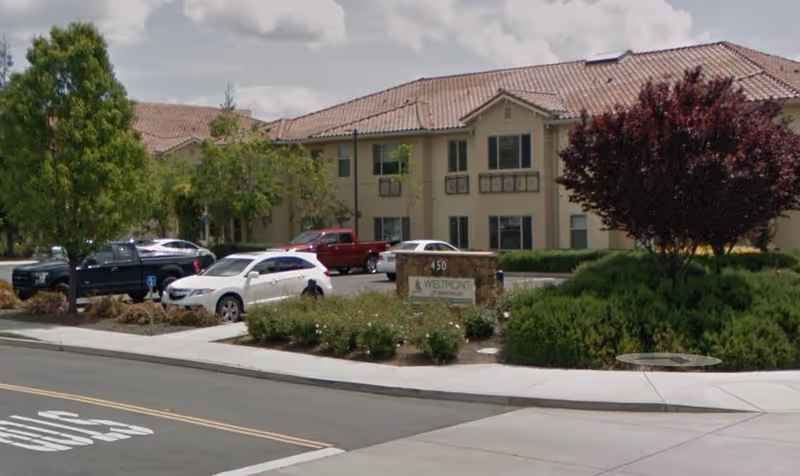 Front view of a two-story beige senior living building with a tiled roof, landscaped entrance, parked cars, and a stone sign reading 'Westmont'.