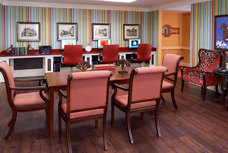 A cozy room with a wooden table surrounded by six upholstered chairs in red fabric. Behind the table, there is a long desk with three red office chairs and three computer monitors. The wall behind the desk features colorful vertical striped wallpaper and framed black and white pictures. To the right, there is a red patterned armchair next to a small side table with a telephone. The floor is dark wood, and a sign on the wall in the background reads 'Library'.