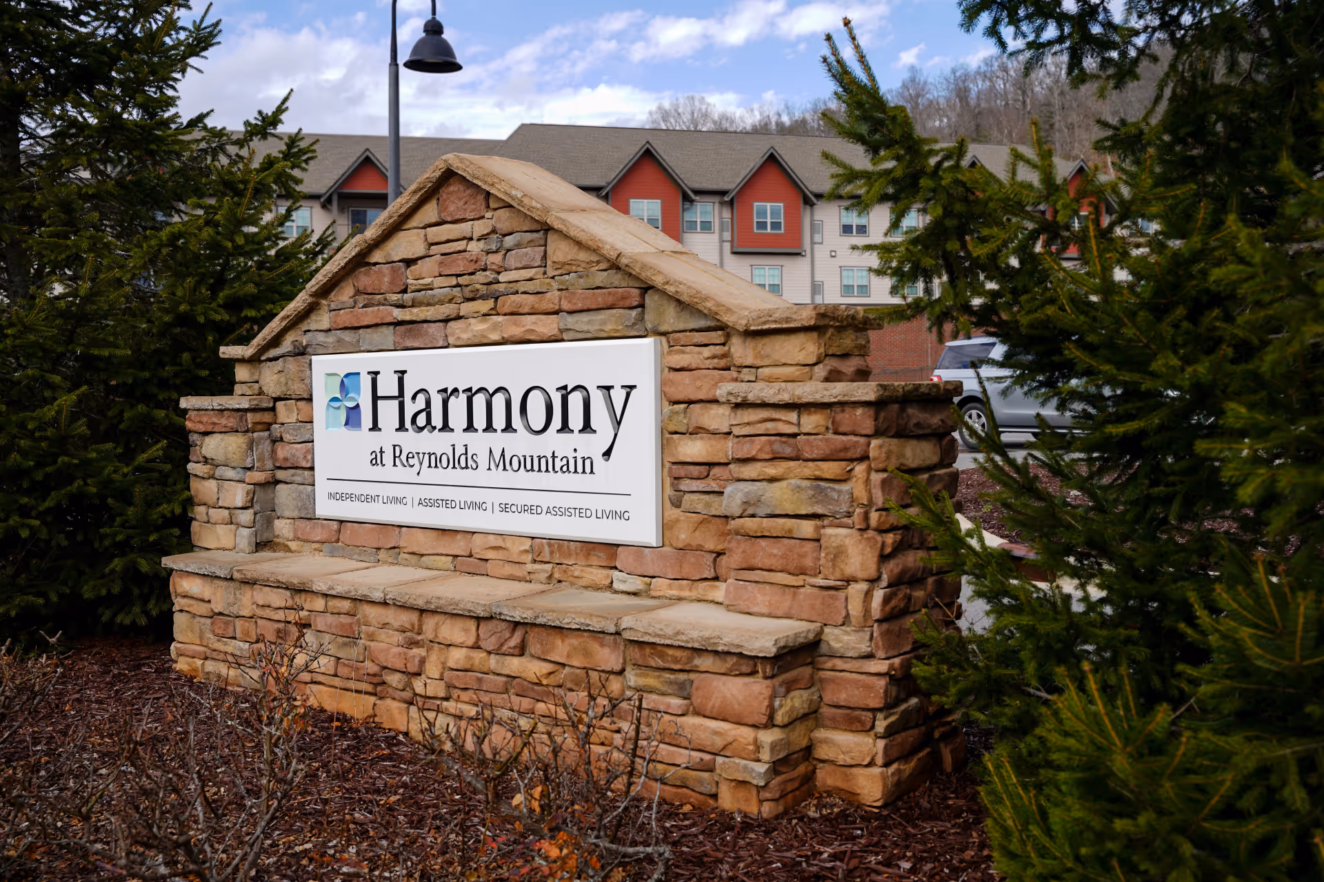 Stone monument sign for Harmony at Reynolds Mountain senior living facility, surrounded by greenery with a building and parked cars in the background under a partly cloudy sky.