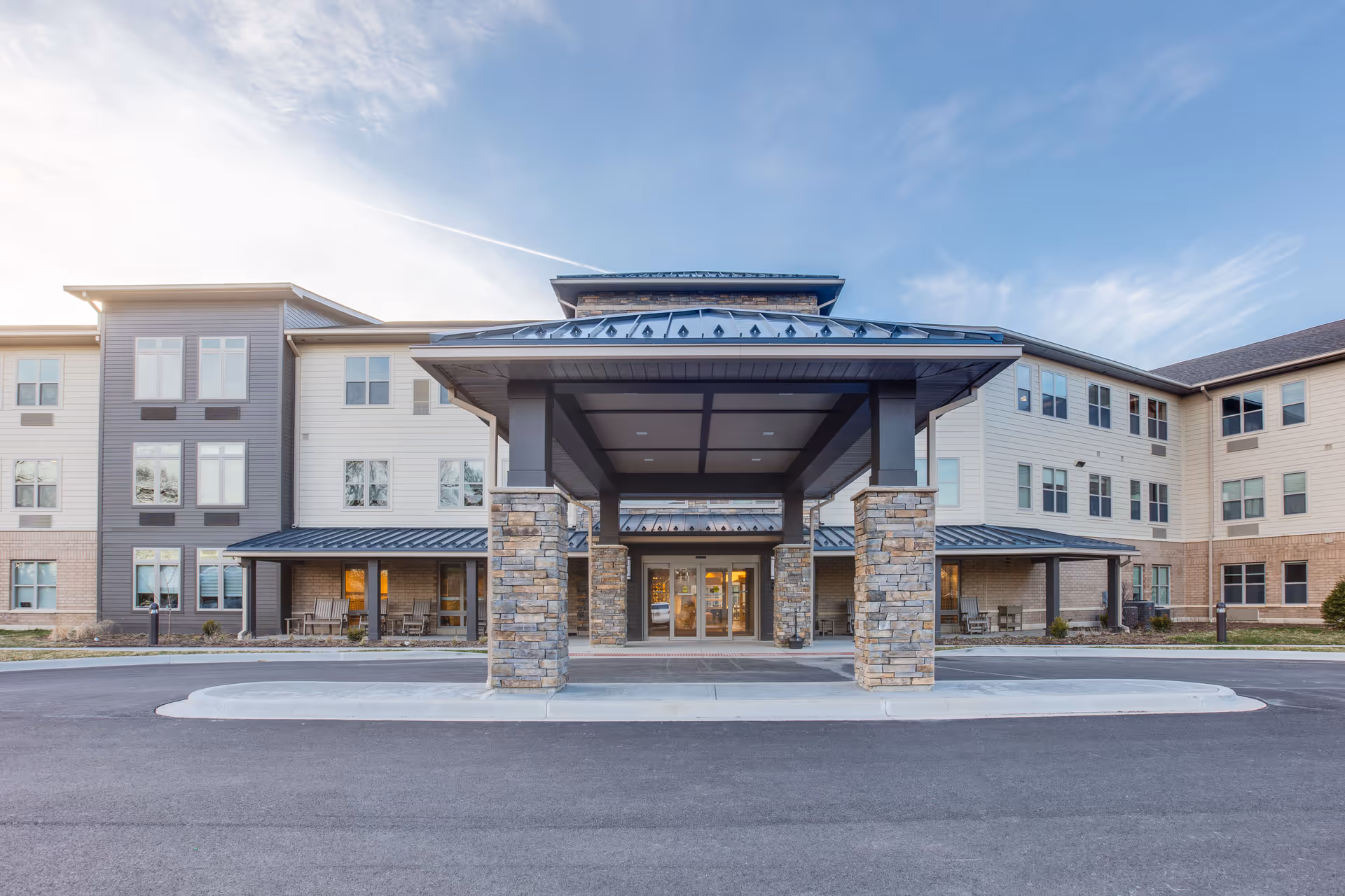 Front exterior view of a modern three-story senior living facility with a covered entrance supported by stone pillars, large windows, and a paved driveway under a partly cloudy sky.