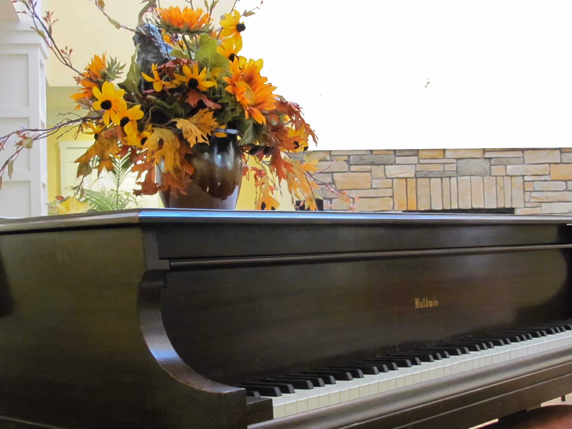 A close-up view of a black Baldwin grand piano with a large vase of yellow and orange flowers placed on top. In the background, there is a stone fireplace and some indoor plants.