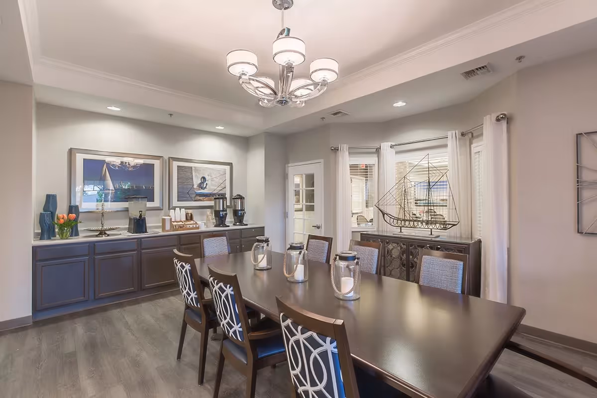 Well-lit dining room with a long dark table and patterned chairs, a buffet with a coffee station and artwork, a chandelier, and a decorative sailboat by the window.