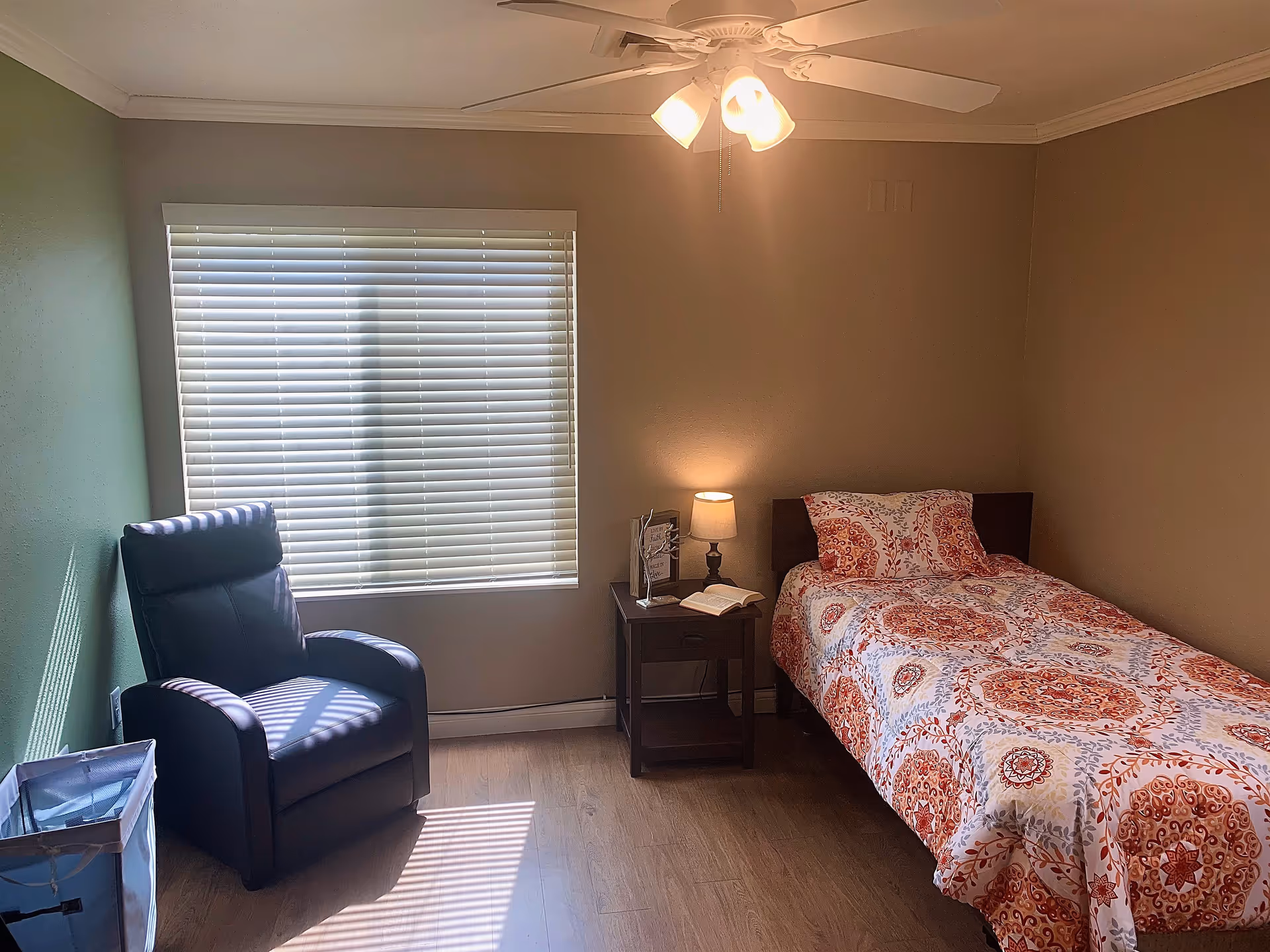 A small bedroom with a single bed covered in a red and white patterned bedspread, a wooden nightstand with a lamp and an open book, a black recliner chair near a window with closed blinds, and a ceiling fan with lights.
