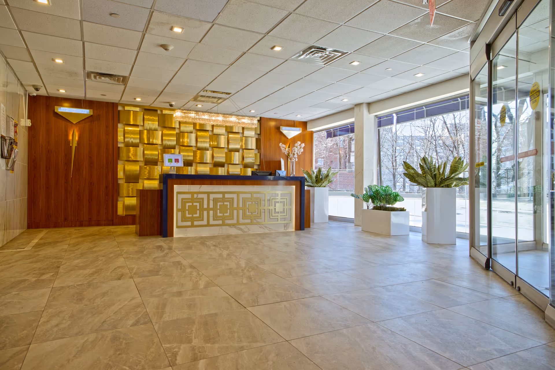 Spacious reception lobby with a decorative gold-accented front desk, large windows, and potted plants.