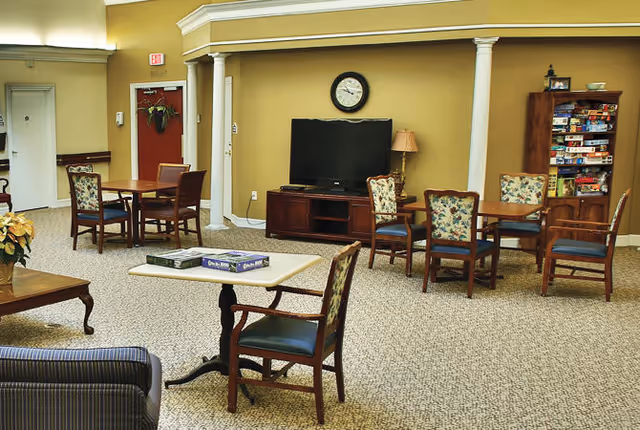 A common area in a senior living facility with several wooden tables and chairs arranged for group activities or games. A television sits on a wooden stand against a beige wall with a clock above it. A tall wooden shelf filled with board games is visible on the right side. The room has carpeted flooring and neutral-colored walls with white columns and trim.