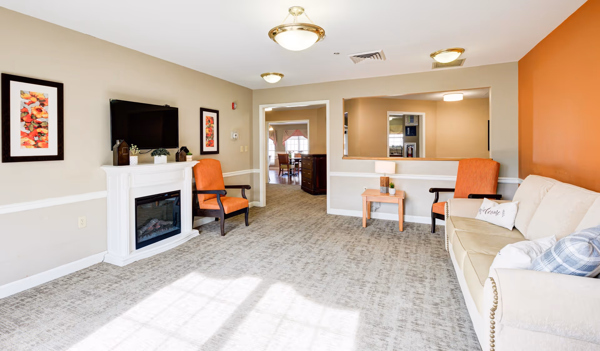 A bright and cozy living room with beige walls and carpeted floor. The room features a white electric fireplace with a flat-screen TV mounted above it, flanked by two framed floral artworks. There are two orange upholstered armchairs with dark wooden arms, a small wooden side table with decorative items, and a beige sofa with patterned pillows. A large window allows natural light to fill the space, and a doorway leads to another room with dining furniture visible.