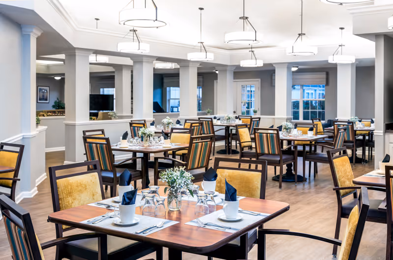 A bright and spacious dining room with multiple wooden tables set for four people each. The chairs have yellow and striped upholstery. Each table is neatly arranged with white plates, cups, silverware, and napkins folded in a standing design. Small floral centerpieces decorate the tables. The room features white walls, large windows, and modern ceiling lights.