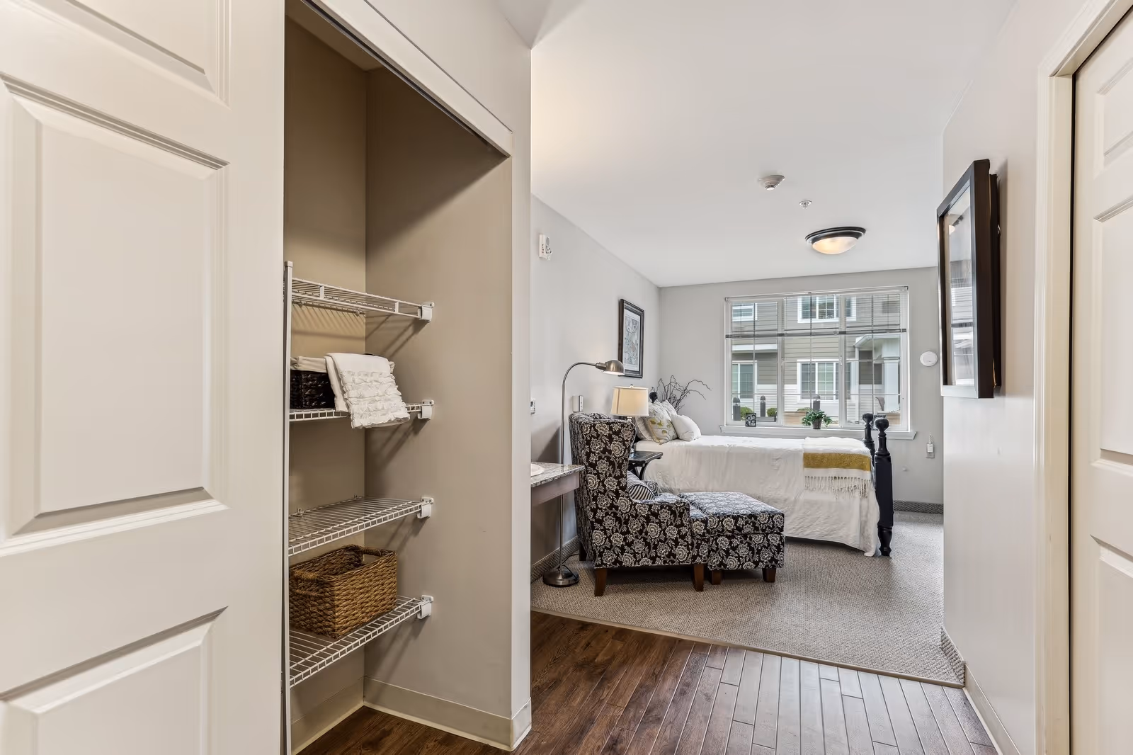 Furnished bedroom with a bed, patterned armchair, window and open closet shelving visible from the entry.