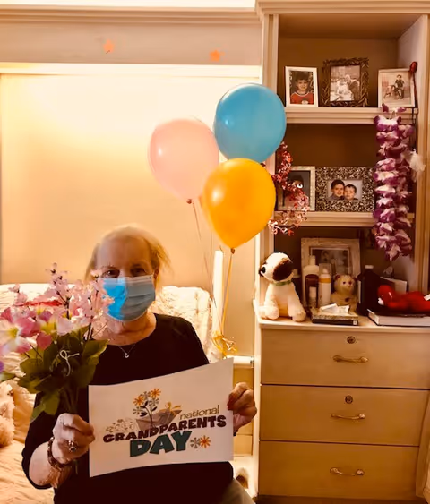 An elderly woman wearing a blue face mask is sitting in a room holding a bouquet of flowers and a sign that reads 'National Grandparents Day'. Behind her are three balloons in pink, blue, and yellow, and a dresser with framed photos, stuffed animals, and decorative items.