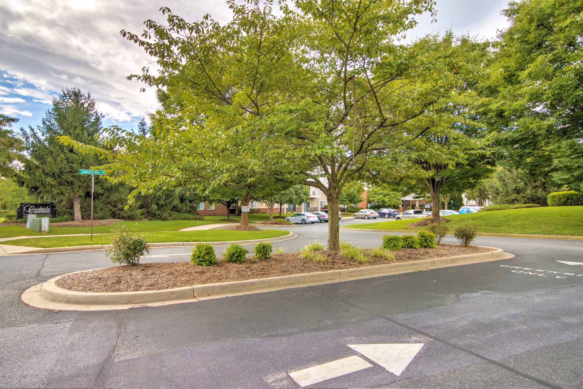Landscaped entrance roundabout with trees, shrubs and parked cars in front of a multi-building senior living complex.