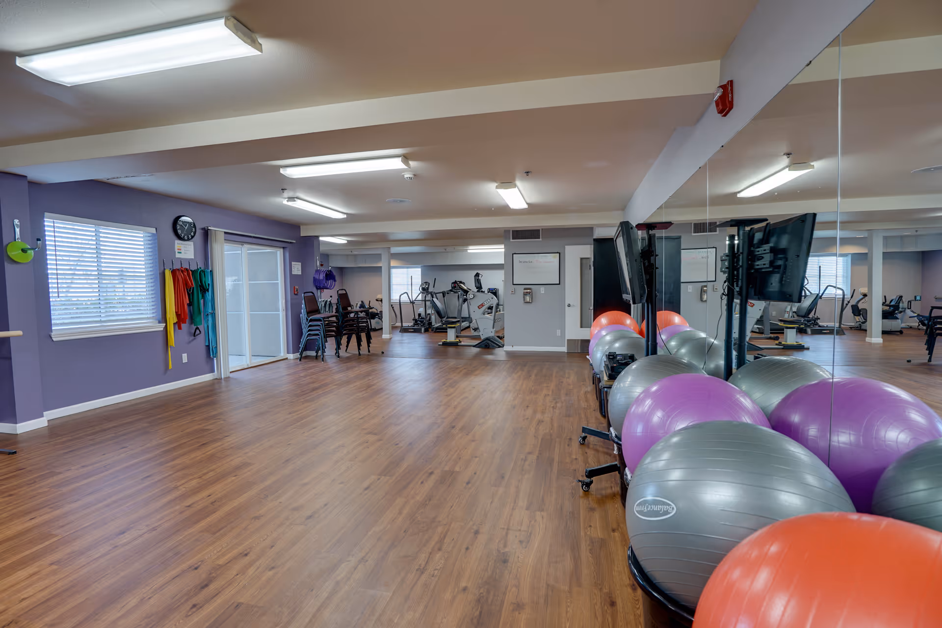 Spacious senior living fitness room with wood floors, exercise balls, cardio machines, mirrored wall and purple accent wall.