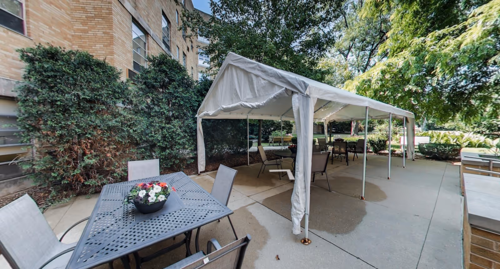 Outdoor patio area at Celebrate Senior Living of Niles with a white canopy tent covering several tables and chairs. There is a metal table with a flower pot in the foreground, surrounded by chairs. The area is surrounded by greenery including bushes and trees, and the building's brick exterior is visible on the left.