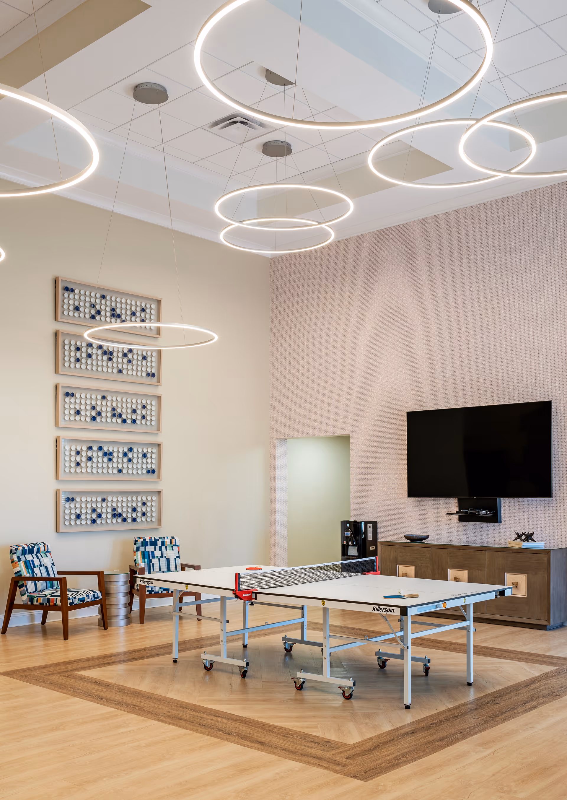 A bright recreational room with a ping pong table in the center, two patterned armchairs against the wall, modern circular ceiling lights, a large flat-screen TV mounted on the wall above a wooden cabinet, and decorative wall art made of ping pong balls.