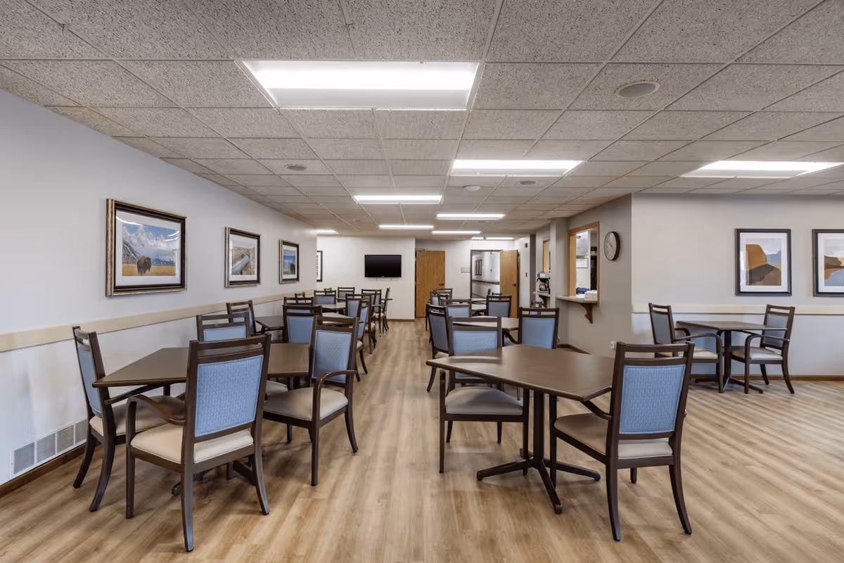 A spacious dining room in a senior living facility with multiple wooden tables and chairs arranged neatly on a light wood floor. The walls are decorated with framed landscape paintings, and there is a wall-mounted television at the far end of the room. The ceiling has recessed lighting panels providing bright illumination.