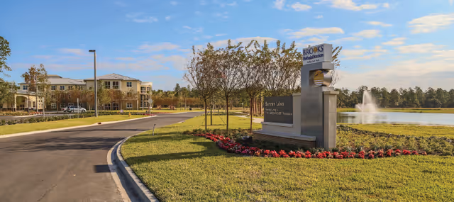 Exterior view of Bartram Lakes Assisted Living Facility - Brooks Rehabilitation showing a curved driveway, landscaped lawn with red flowers, trees, a large sign with the facility name, and a water fountain in a pond under a blue sky with scattered clouds.